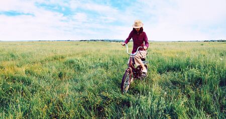 Hipster teenager girl in straw hat cycling on green meadow in summerの写真素材