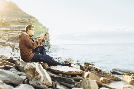 Traveler hipster young man takes photographs with vintage photo camera on coastline near the sea. Image with sunlight effectの写真素材