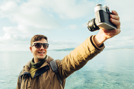 Traveler happy young man takes photographs self portrait with old photo camera on coastline on background of seaの写真素材