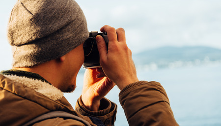 Traveler hipster guy takes photographs with vintage photo camera on coastline near the seaの写真素材
