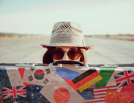 Hipster girl in a hat and sunglasses looks out from vintage suitcase. Suitcase with stamps flags of different countryの写真素材