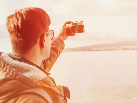 Traveler young man doing selfie with old photo camera on coastline. Image with sunlight effect. Focus on cameraの写真素材