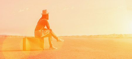 Traveler woman sits on a suitcase and looks away on road. Image with sunlight effectの写真素材