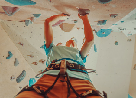 Girl in safety harness climbing artificial boulder indoors, view from belowの写真素材