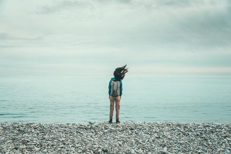 Traveler girl with backpack standing on coast near the sea in windy weather in the evening, her hair fluttering in the windの写真素材