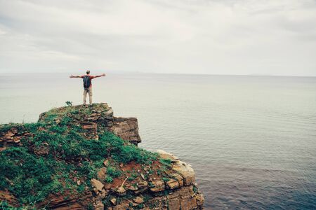 Happy traveler man standing  with raised arms on peak of rock and enjoying view of seaの写真素材