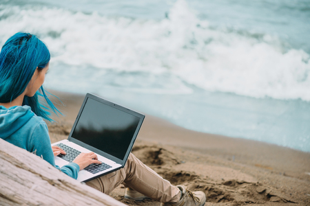 Freelancer young woman with blue hair working on laptop on beach near the sea. Freelance concept. Focus on handの写真素材
