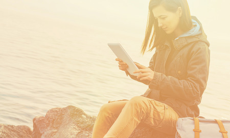 Traveler girl sitting on coast near the sea and working on digital tablet. Image with sunlight effectの写真素材