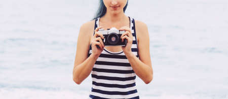 Young woman in striped blue shirt standing with vintage photo camera on background of sea in summer