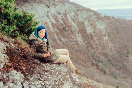 Smiling young woman wearing parka sitting in the mountains and looking at camera. Space for text in...