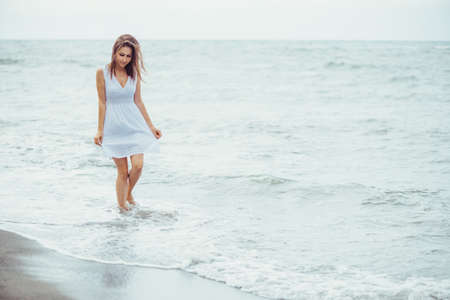 Beautiful young woman in white dress walking on sea coast in summerの写真素材