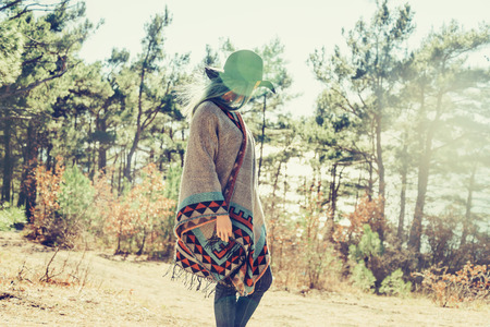 Fashionable girl in hat and poncho walking in forest among pine trees at sunny dayの写真素材