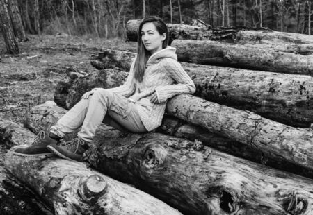 Young woman sitting on stack of tree trunk, girl resting outdoor. Black and white imageの写真素材