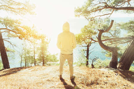 Young man standing on coast in the forest among pine trees and looking at sea in sunny day, rear viewの写真素材