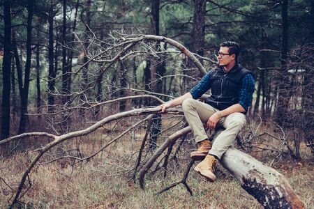 Explorer young man sitting on fallen tree in the forest. Traveler man resting outdoorの写真素材