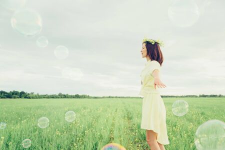 Happy beautiful young woman in flower wreath walking on summer meadow among soap bubblesの写真素材