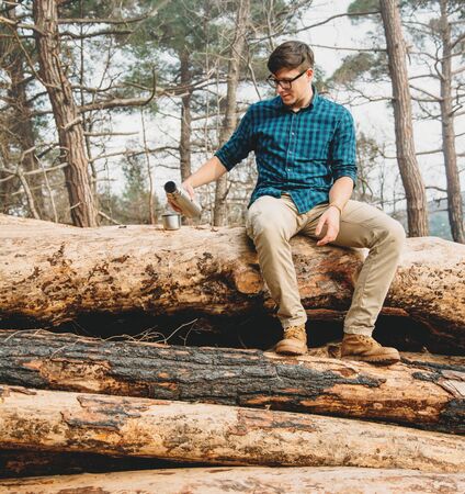 Traveler young man sitting on tree trunk and pouring tea from thermos to cup in the forest. Man has a picnic outdoorの写真素材