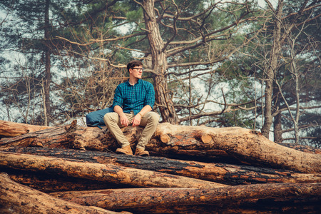 Explorer traveler young man with backpack sitting on felled tree trunk in the pine forest outdoor in summerの写真素材