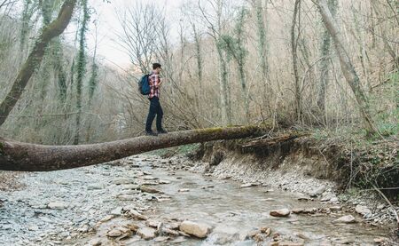 Hiker young man with backpack crossing river on fallen tree trunk in the forestの写真素材
