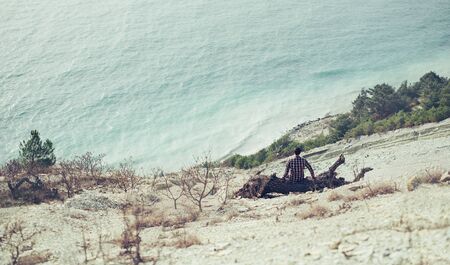 Young man sitting on snag on coast and enjoying view of sea, rear viewの写真素材