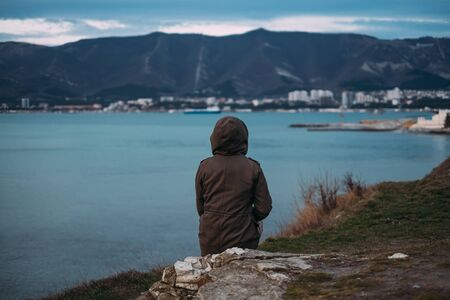 Unrecognizable woman sitting on coast in parka jacket with hood and enjoying view of mountain and...