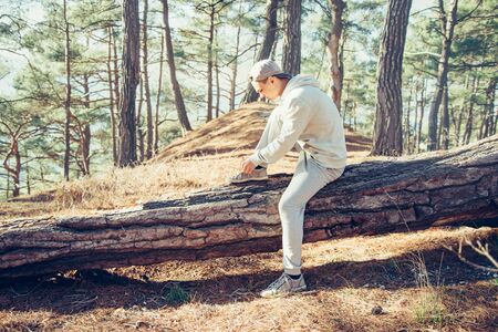 Sporty young man sitting on fallen tree trunk and tying shoelaces outdoor in the pine forestの写真素材