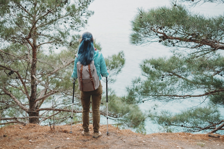 Hiker young woman with trekking poles and backpack standing on steep coast among pine trees and looking at sea, rear viewの写真素材