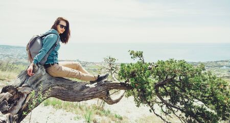 Beautiful traveler young woman with backpack resting on juniper tree in summer outdoor