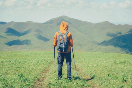 Unrecognizable hiker young man with backpack and trekking poles walking on path in the mountains in summer outdoor, rear viewの写真素材