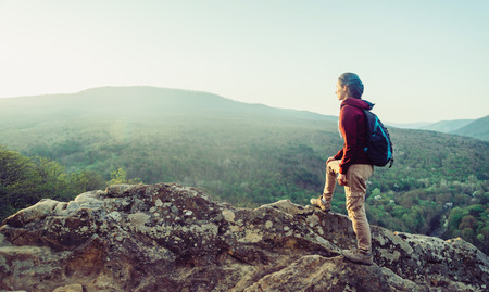 Hiker young woman with backpack standing on peak of rock in the mountains and looking into the...