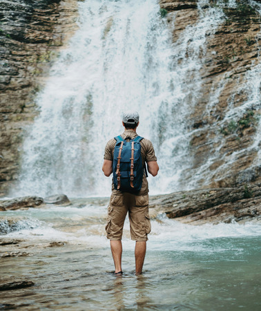 Unrecognizable traveler young man with backpack standing in river and looking at waterfall in summer outdoor, rear viewの写真素材