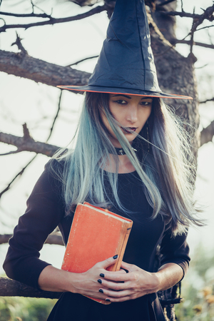 Dark young witch standing with book in the forest, looking at camera. Space for text on bookの写真素材