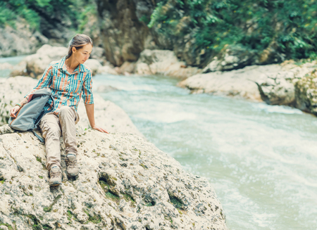 Traveler young woman with backpack sitting on stone and looking on mountain river in summerの写真素材