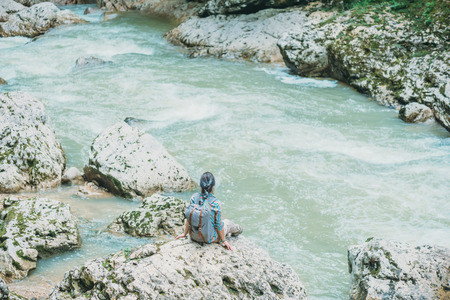 Traveler young woman with backpack sitting on rocky stone and looking on mountain river in summerの写真素材