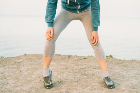 Fitness young woman warming-up her knee and preparing to run on coast in summerの写真素材
