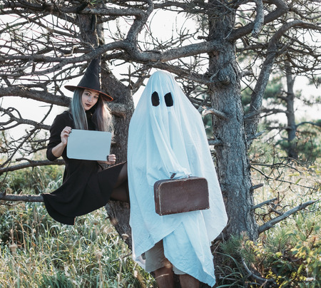 Halloween couple. White ghost and beautiful witch with empty poster standing near old dry trees in the forestの写真素材