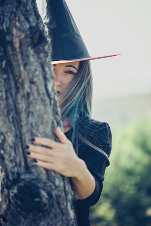 Young woman in costume of witch looks out from behind a treeの写真素材