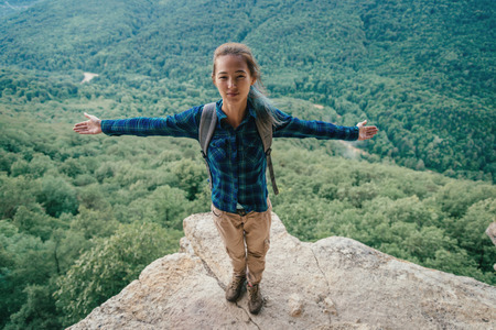 Happy hiker young woman with backpack standing with raised arms in summer mountains, looking at cameraの写真素材