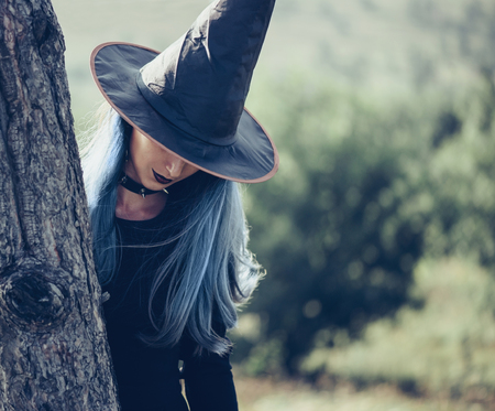 Beautiful mysterious young witch in black hat and dress standing near a tree. Halloween themeの写真素材