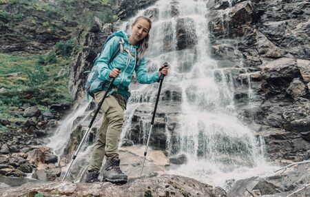 Smiling backpacker young woman with trekking poles standing near the waterfallの写真素材