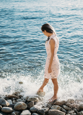 Beautiful young woman walking on pebble beach near the sea, looking at sea wavesの写真素材