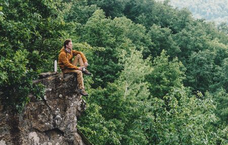 Traveler young man resting on top of cliff and drinking tea from flask outdoorの写真素材