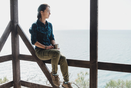 Casual style young woman sitting on wooden terrace with cup of tea on background of sea and enjoying view of natureの写真素材