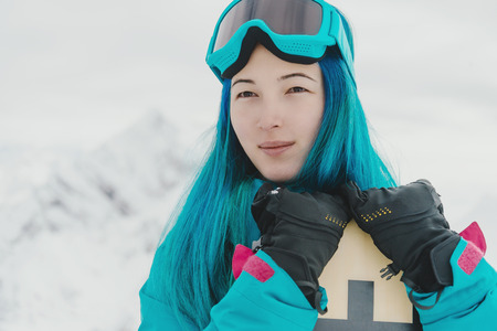 Smiling young woman with blue hair standing with snowboard on background of snowy mountainsの写真素材