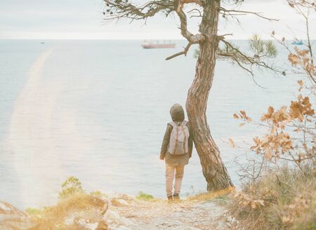 Unrecognizable female traveler with backpack standing near a tree and looking at sea in sunny day, rear view. Image with sunlight effect.の写真素材