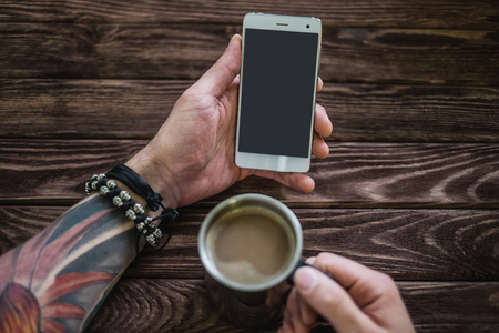 Young man sitting with a smartphone and a cup of coffee at a wooden table, point of view. Free space on screen of phone.の写真素材