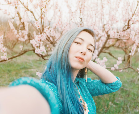 Beautiful young woman with blue hair making selfie in blooming spring garden.の写真素材