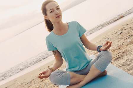 Young woman meditating in pose of lotus on sand shore on background of sea, looking at camera.の写真素材
