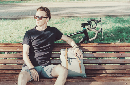 Handsome guy cyclist sitting with backpack on wooden bench in summer park.の写真素材
