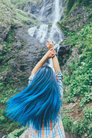 Tourist young woman with blue hair standing with raised arms in front of waterfall, concept of travel and freedom.の写真素材